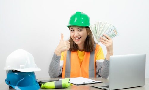 Ingeniera civil con chaleco naranja y casco verde sosteniendo billetes en su mano, está sentada en un escritorio