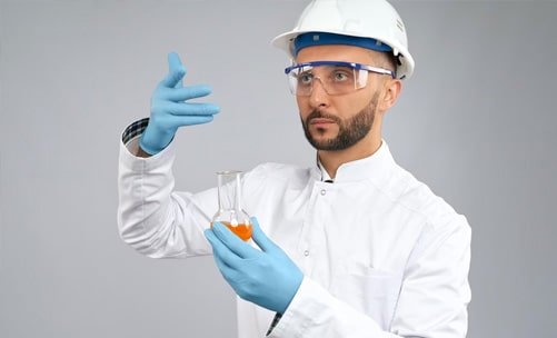 Ingeniero civil laboratorista vistiendo bata y casco blancos, guantes plásticos azules y gafas transparentes de seguridad, revisando una muestra que está en un matraz.