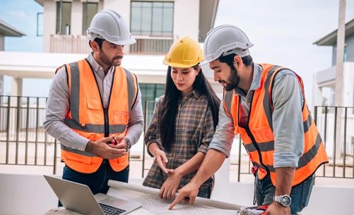 Tres ingenieros civiles revisan los planos de una construcción sobre una mesa, llevan puesto su equipo de seguridad.
